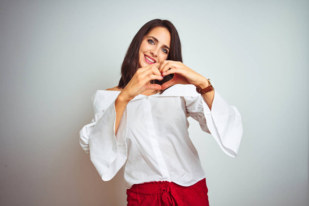 Молодая красивая женщина в футболке стоит на белом изолированном фоне улыбаясь в любви показывая символ сердца и форму с руками. Романтическая концепция
. - Фото, изображение