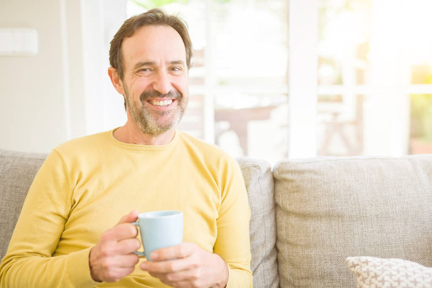 Knappe middelbare leeftijd man zit op de Bank ontspannen een kopje koffie drinken en glimlachend in de camera thuis - Foto, afbeelding