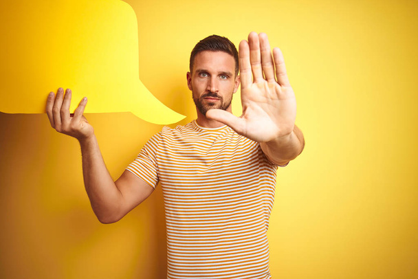 Молодой человек держит пустой пузырь речи для сообщения на желтом изолированном фоне с открытой рукой делает знак стоп с серьезным и уверенным выражением, защитный жест
 - Фото, изображение