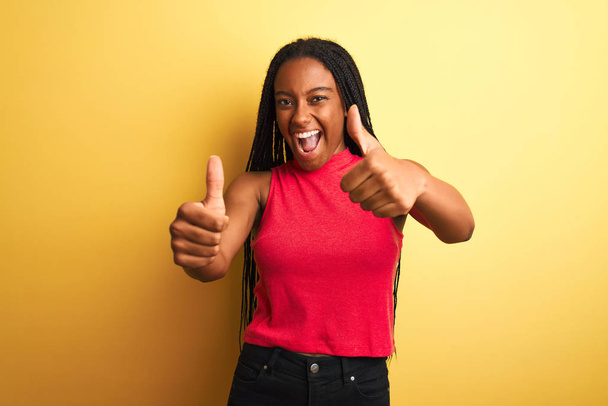 African american woman wearing red casual t-shirt standing over isolated yellow background approving doing positive gesture with hand, thumbs up smiling and happy for success. Winner gesture. - Фото, зображення