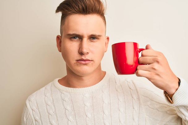 Молодой красивый мужчина пьет красную чашку кофе стоя на изолированном белом фоне с уверенным выражением на умном лице, думая серьезно
 - Фото, изображение