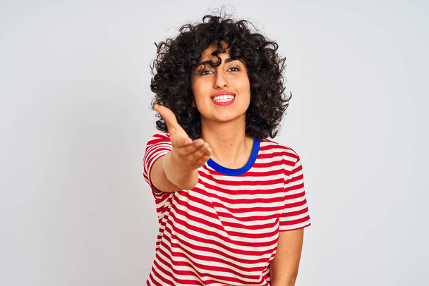Mujer árabe joven con el pelo rizado con camiseta a rayas sobre fondo blanco aislado sonriendo amistoso ofreciendo apretón de manos como saludo y bienvenida. Negocios exitosos
. - Foto, Imagen
