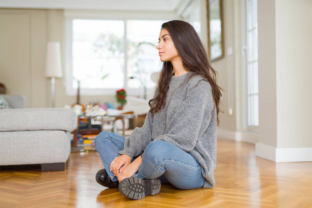 Young beautiful woman sitting on the floor at home looking to side, relax profile pose with natural face with confident smile. - Foto, Imagen