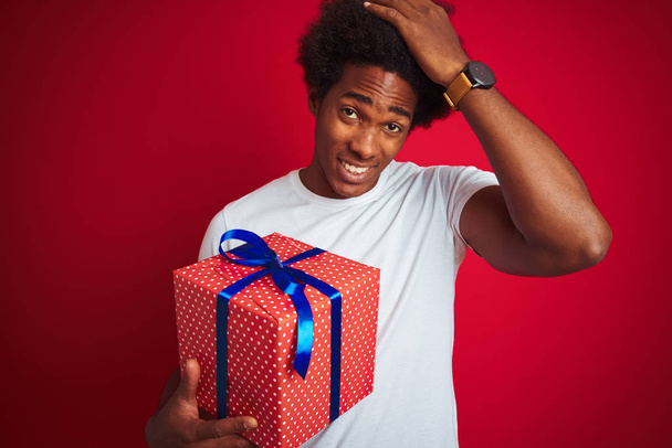 Mladý afroamerický Američan, který drží narozeninový dar stojící nad izolovaným červeným pozadím, zdůrazňoval rukou na hlavě, šokován hanbou a překvapením tváře, rozzlobený a frustrovaný. Strach a rozčílený omyl. - Fotografie, Obrázek