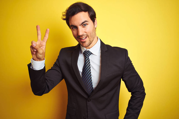 Молодой привлекательный бизнесмен в костюме и галстуке стоит на изолированном желтом фоне и улыбается, глядя в камеру, показывая пальцы, делающие знак победы. Номер два:
. - Фото, изображение