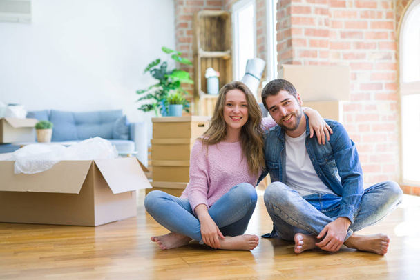 Νεαρό όμορφο ζευγάρι που κινείται σε ένα νέο σπίτι καθισμένος στο πάτωμα με ένα χαρούμενο και δροσερό χαμόγελο στο πρόσωπο. Τυχερός άνθρωπος. - Φωτογραφία, εικόνα