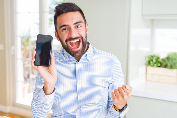 Handsome hispanic business man showing smartphone screen screaming proud and celebrating victory and success very excited, cheering emotion - 写真・画像