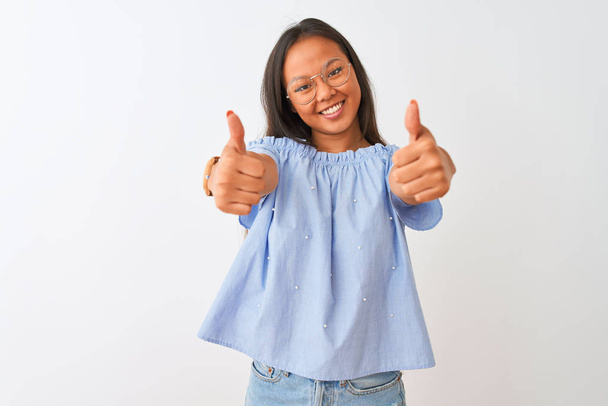 Jeune Chinoise portant un t-shirt bleu et des lunettes sur fond blanc isolé approuvant faire un geste positif avec la main, le pouce levé souriant et heureux pour le succès. Le geste du gagnant
. - Photo, image