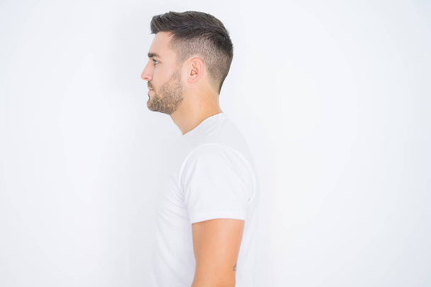 Jonge knappe man dragen casual wit t-shirt over witte geïsoleerde achtergrond op zoek naar kant, ontspannen profiel pose met natuurlijk gezicht met zelfverzekerde glimlach. - Foto, afbeelding