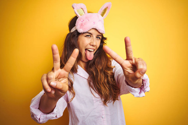junge Frau in Pyjama und Schlafmaske vor gelbem Hintergrund lächelnd mit herausgestreckter Zunge, die Finger beider Hände beim Siegeszeichen zeigt. Nummer zwei. - Foto, Bild