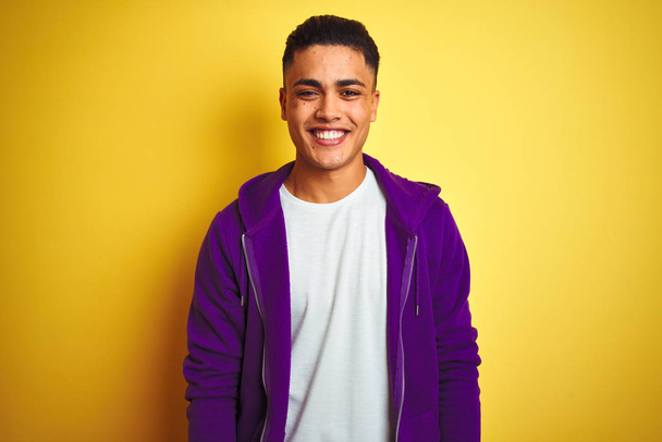 紫色のスウェットシャツを着た若いブラジル人男性が、黄色の背景に立ち、幸せでクールな笑顔を浮かべていた。ラッキーな人. - 写真・画像