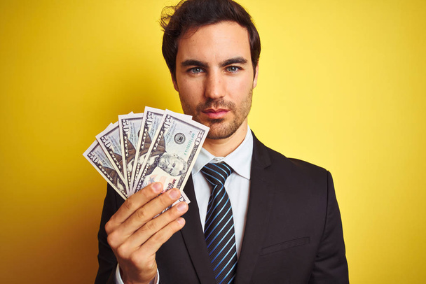 Jeune homme d'affaires beau portant costume tenant des dollars sur fond jaune isolé avec une expression confiante sur la pensée intelligente visage grave
 - Photo, image