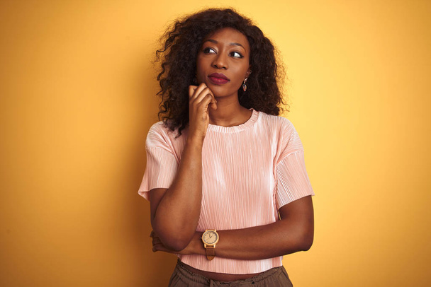 African American kobieta ubrana różowy t-shirt stojący na białym tle żółtym z ręki na podbródek myślenia o pytanie, pokutne wyrażenie. Uśmiechnięta z przemyślaną twarzą. Koncepcja wątpliwości. - Zdjęcie, obraz