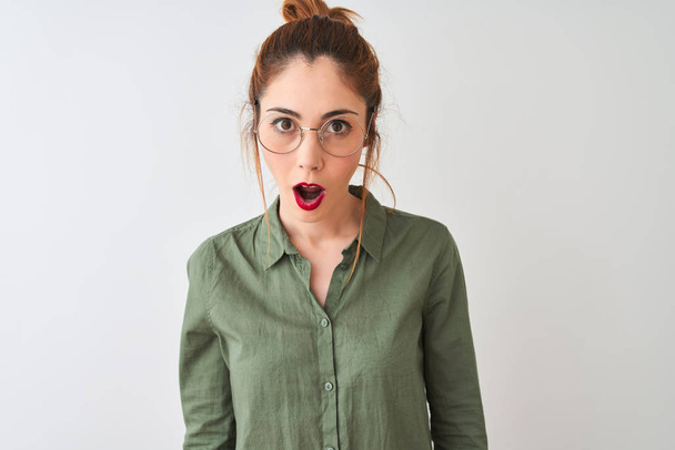 Redhead vrouw dragen groen shirt en glazen staande over geïsoleerde witte achtergrond bang en geschokt met verrassing expressie, angst en opgewonden gezicht. - Foto, afbeelding