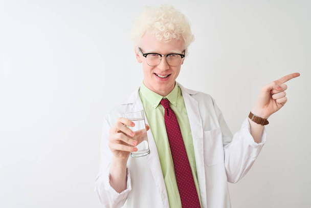 孤立した白い背景の上に水のガラスを持つ眼鏡をかけたアルビノの科学者の男は、手と指を横に指差して非常に幸せ - 写真・画像