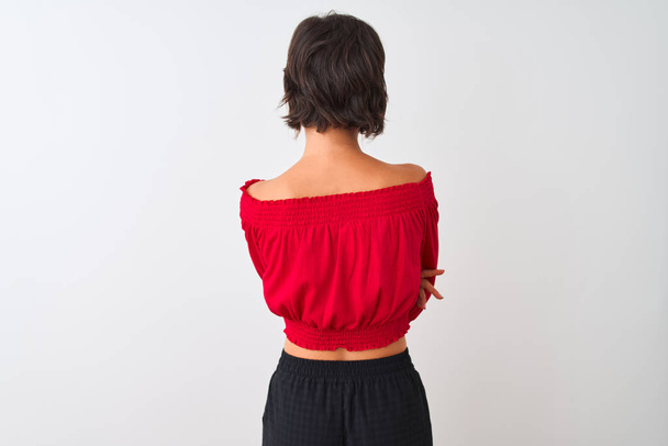 赤い夏のTシャツを着た若い美しい女性は、交差した腕で後ろ向きに立って孤立した白い背景の上に立っている - 写真・画像