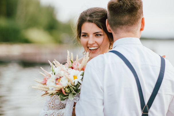 Ευτυχισμένο νιόπαντρο ζευγάρι, χαμογελαστή νύφη μελαχρινή νεαρή γυναίκα με το στυλ Μπόχο ανθοδέσμη με τον γαμπρό, κοντινό πορτρέτο σε εξωτερικούς χώρους - Φωτογραφία, εικόνα
