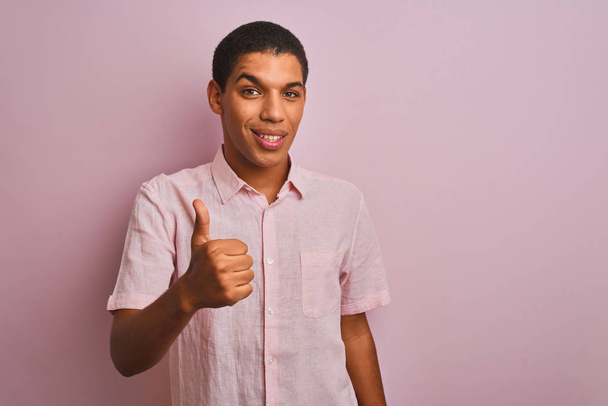 junger gutaussehender arabischer Mann in lässigem Hemd, der vor isoliertem rosa Hintergrund steht und mit der Hand fröhlich Daumen nach oben gestikuliert. Zustimmender Blick in die Kamera zeigt Erfolg. - Foto, Bild