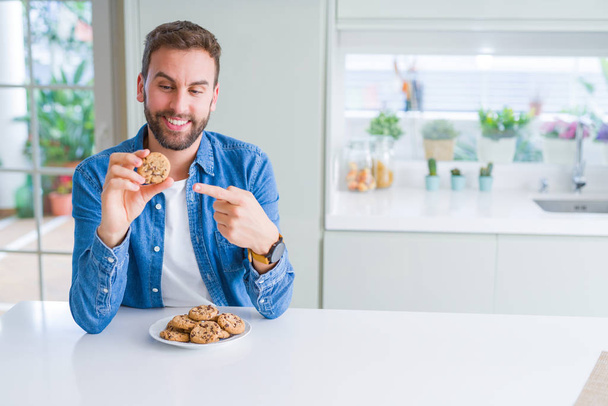 Handsome man eating chocolate chips cookies very happy pointing with hand and finger - Photo, Image