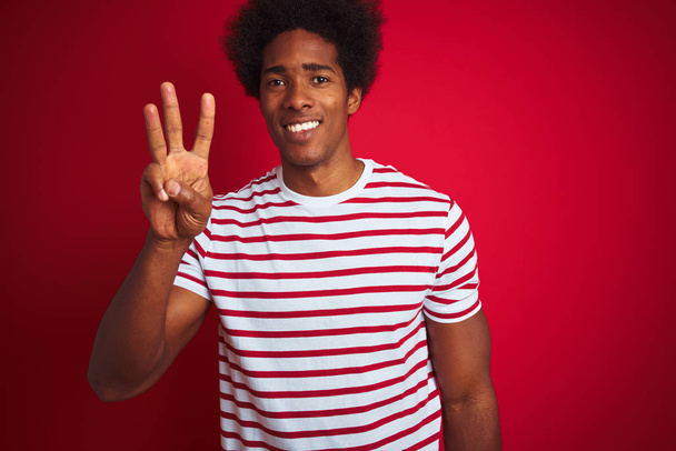 Young african american man with afro hair wearing striped t-shirt over isolated red background showing and pointing up with fingers number three while smiling confident and happy. - Photo, Image