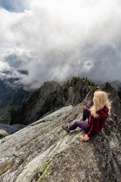 Adventurous Girl on top of a rugged rocky mountain during a cloudy summer morning. Taken on Crown Mountain, North Vancouver, BC, Canada. - Foto, Imagen