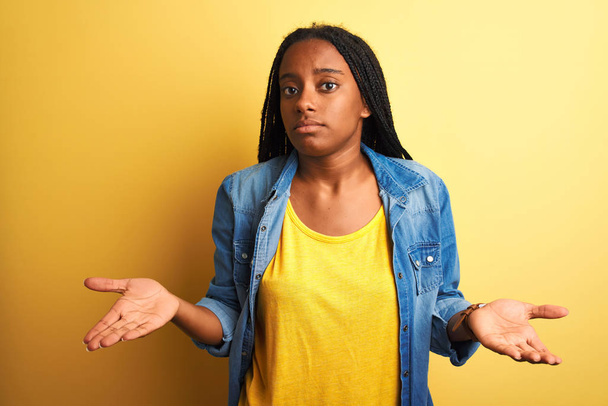 Młoda Afroamerykanka ubrana w dżinsową koszulę stojąca nad odizolowanym żółtym tłem, nieświadoma i zdezorientowana z otwartymi ramionami.. - Zdjęcie, obraz