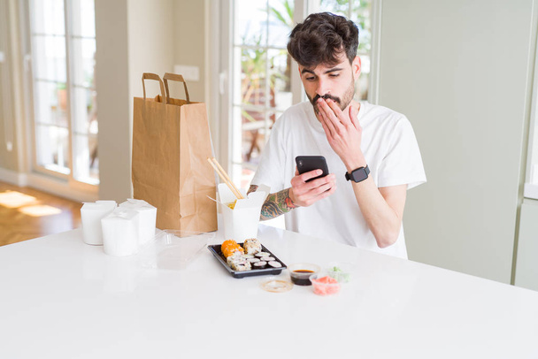 Jeune homme manger des sushis asiatiques de la livraison à domicile et de commander de la nourriture en utilisant smartphone app couvrir bouche avec la main choquée par la honte pour erreur, expression de la peur, peur dans le silence, concept secret
 - Photo, image