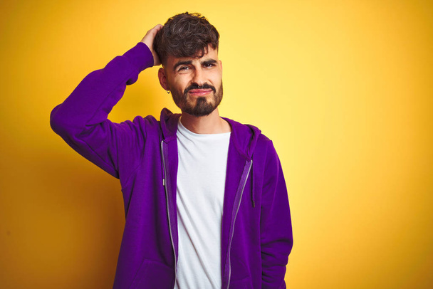 Young man with tattoo wearing sport purple sweatshirt over isolated yellow background confuse and wonder about question. Uncertain with doubt, thinking with hand on head. Pensive concept. - Photo, Image