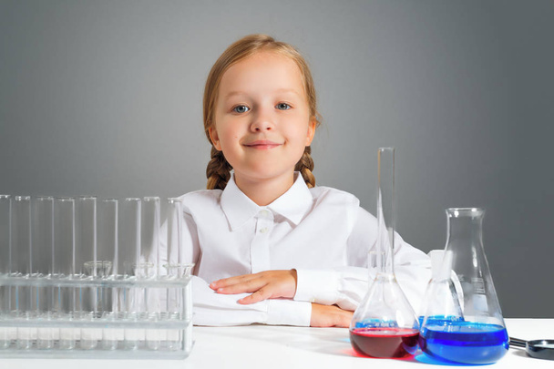 Mała dziewczynka z kolb do chemii siedzi przy stole i patrzy na aparat na szarym tle. - Zdjęcie, obraz