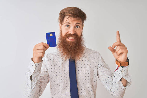 Młody rudy irlandzki biznesmen posiadający kartę kredytową stojący na białym tle bardzo szczęśliwy wskazując ręką i palcem na bok - Zdjęcie, obraz