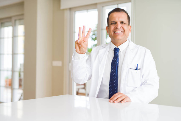 Keski-ikäinen lääkäri mies yllään lääketieteellinen takki klinikalla näyttää ja osoittaa sormilla numero kolme hymyillen itsevarma ja onnellinen
. - Valokuva, kuva