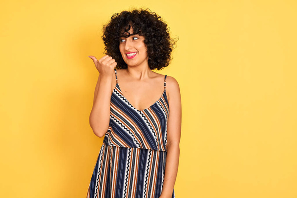 Arab woman with curly hair wearing striped colorful dress over isolated yellow background smiling with happy face looking and pointing to the side with thumb up. - Valokuva, kuva
