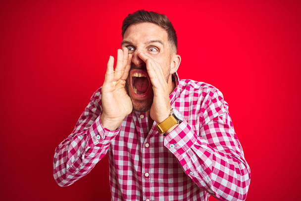 Jonge knappe man over rode geïsoleerde achtergrond schreeuwen boos uit luid met handen over de mond - Foto, afbeelding