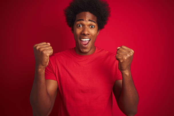 Young american man with afro hair wearing t-shirt standing over isolated red background celebrating surprised and amazed for success with arms raised and open eyes. Winner concept. - Fotó, kép