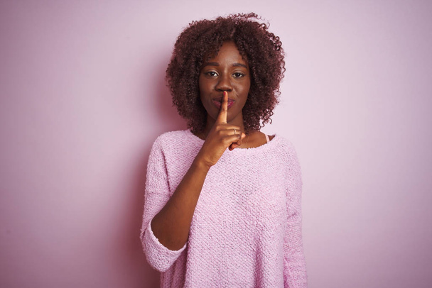 Mladá Africká Afro žena nosí svetr, který stojí na osamělém růžovém pozadí a žádá, aby byl zticha prstem na rtech. Ticho a tajný koncept. - Fotografie, Obrázek
