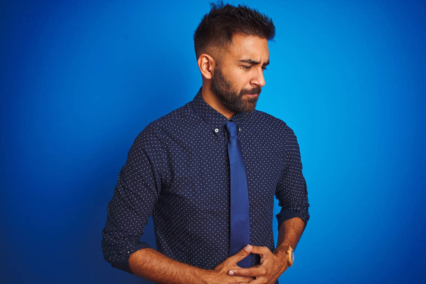Jeune homme d'affaires indien portant chemise élégante et cravate debout sur fond bleu isolé avec la main sur le ventre parce que les nausées, la maladie douloureuse se sentent mal. Concept Ache
. - Photo, image