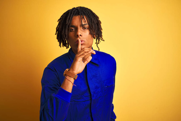 Afro worker man with dreadlocks wearing mechanic uniform over isolated yellow background asking to be quiet with finger on lips. Silence and secret concept. - 写真・画像