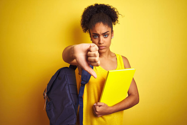 Americká studentka v batohu drží zápisník na izolovaném žlutém pozadí s rozzlobenou tváří, záporné znamení ukazující nechuť k palci, koncept odmítnutí - Fotografie, Obrázek
