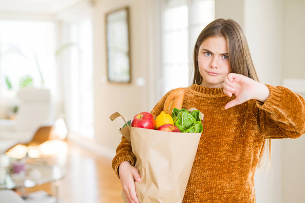 Beautiful young girl holding paper bag of fresh groceries with angry face, negative sign showing dislike with thumbs down, rejection concept - 写真・画像