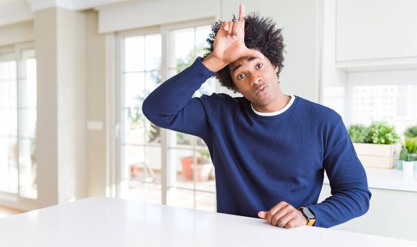 Nuori afrikkalainen amerikkalainen mies yllään rento villapaita istuu kotona pilkkaamassa ihmisiä, joilla on sormet otsassa tekemässä luuseri ele pilkkaa ja loukkaavaa
. - Valokuva, kuva