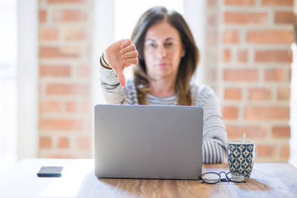 怒った顔をしたコンピュータラップトップを使用して自宅で仕事をしている中年のシニア女性、親指を下にして嫌いを示す否定的なサイン、拒絶概念 - 写真・画像