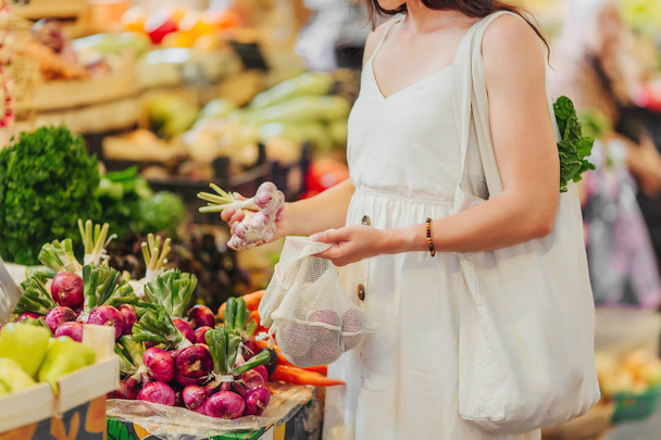 Young Woman puts fruits and vegetables in cotton produce bag at food market. Reusable eco bag for shopping. Zero waste concept. - Photo, Image