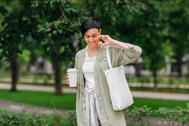 Woman with short hair holding reusable coffee cup and eco bag enjoying morning. Eco friendly concept. - Foto, imagen
