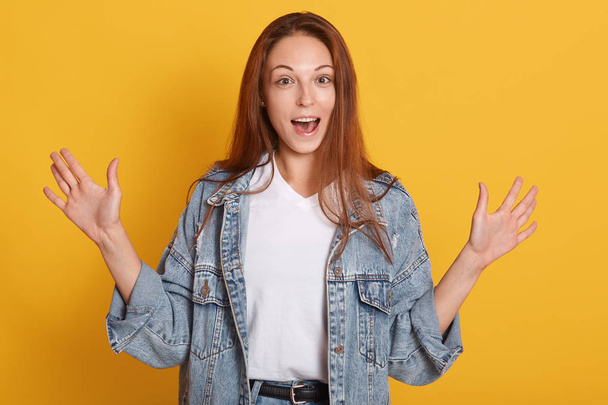 Portrait of excited young woman dressed denim jacket and white t shirt, looking camera and spreading hands, keeping mouth open isolated on yellow background in studio. People emotions concept. - Valokuva, kuva