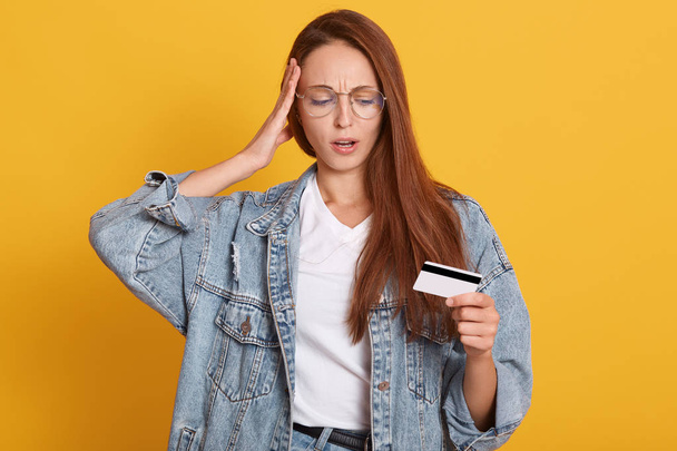 Portrait of astonished young womandressed denim jacket and white shirt, keeping mouth opened, putting hand on head, hold credit bank card and looks on it, posing isolated on yellow background. - 写真・画像