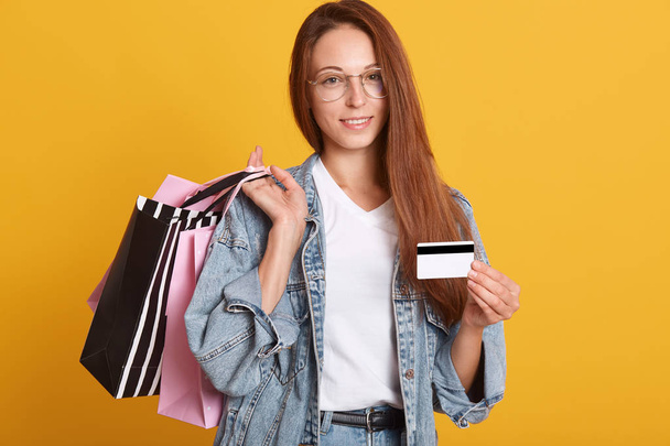 Close up portrait of beautiful stylish young woman with dark hair, dressed demin jacket, white t shirt and spectacles, holding shopping bags and credit card, looking at camera with charming smile. - Foto, afbeelding