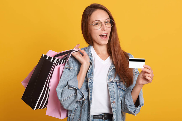 Indoor photo of beautiful smiling Caucasian woman with shopping bags in hands and shows credit card, studio shot isolated over yellow background, wears denim jacket and glasses. Online shopping cocept - Foto, afbeelding