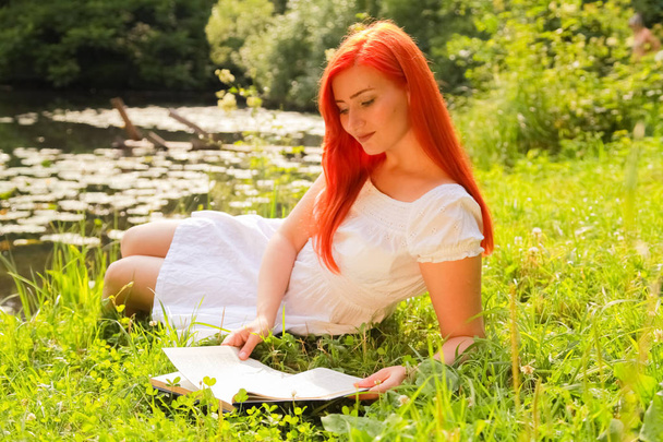 девочка-подросток с рыжими волосами в белом хлопчатобумажном платье отдохнуть и читать книгу в летнем жарком городском парке
 - Фото, изображение