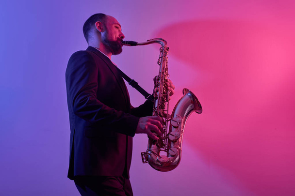 Portrait of professional musician saxophonist man in suit plays jazz music on saxophone, pink background in a photo studio, side view - Photo, Image
