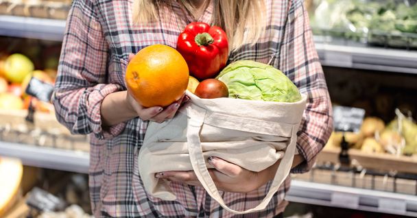 Молода дівчина тримає фрукти та овочі в еко сумці на фоні магазину
 - Фото, зображення